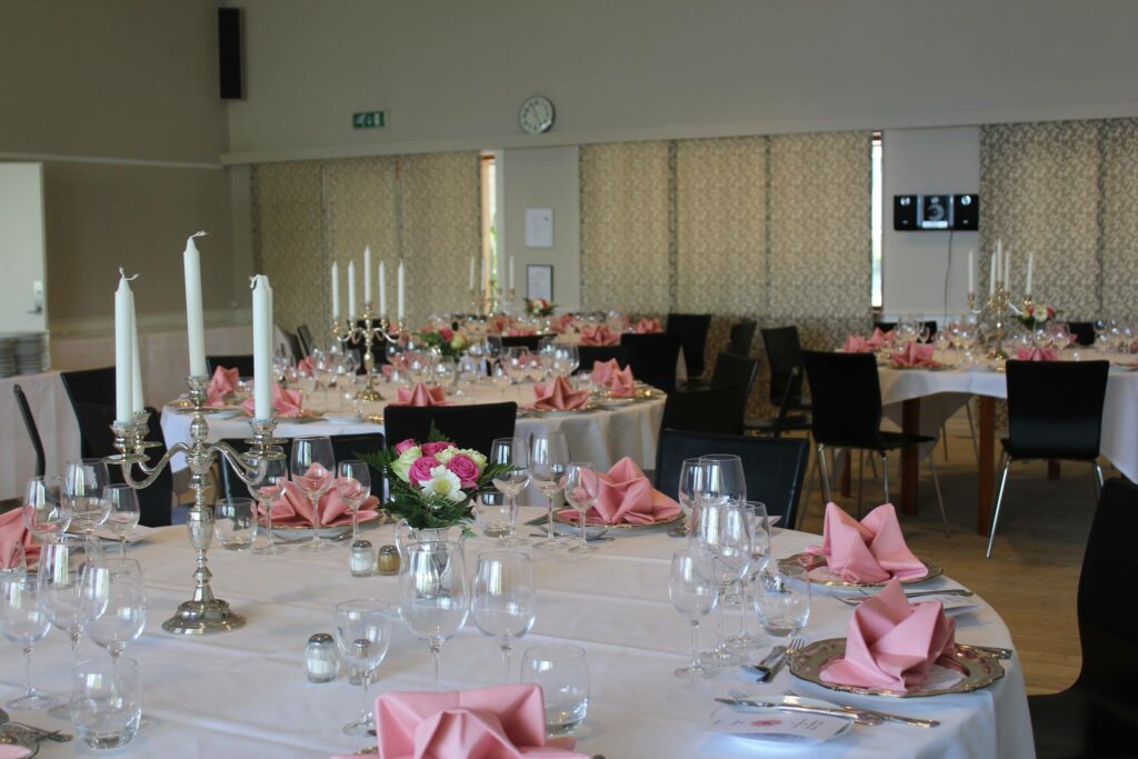 Festsal dækket op med runde borde, hvide duge og lyserøde servietter
